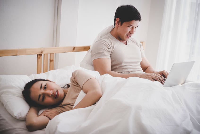 Stock photo of woman, man in bed. He's watching something on laptop, she's frustrated, angry.