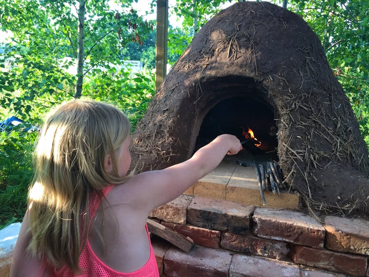 John Scott, of Woodville, built a cob oven in his backyard from potter’s clay, straw and bricks. It is part of his plan to better understand and appreciate where his food comes from. Here, his daughter tends to the fire.