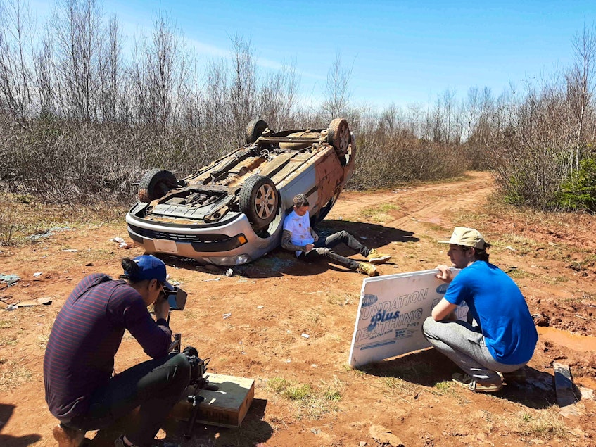 Greg MacDonald (Problematic) sits by a rolled-over car as members of the production team look on during a recent shoot for his latest video.
 - Contributed