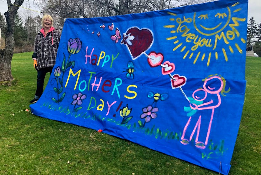 Barb Chiasson stands next to the Mother's Day banner she painted and hung outside her Coxheath home. While the tattoo artist has the sign on display for all mothers, she made it for her mother who lives in Ontario. Chiasson stood with her head peeking over the sun in the photo for her mom. "Oh, she cried," she said with a smile on Friday when asked if she'd pose for a picture with her latest artwork. Along with the Mother's Day sign, Chiasson puts up decorations for all holidays including Easter, Halloween and Christmas. For the anniversary of the mass shooting in Portapique, N.S., Chiasson put signs on the telephone poles near her home which read, "All you need is love" — the popular song by The Beatles. NICOLE SULLIVAN/CAPE BRETON POST 