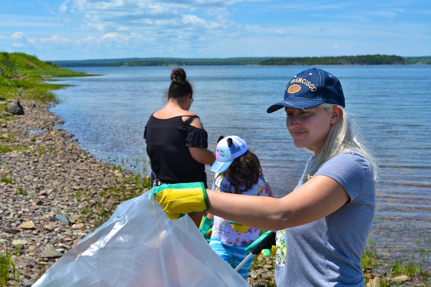 Karina Doucette holds the garbage bag while Oksana Doucette and Maxine Doucette pick up garbage on the shore in Potlotek First Nation. Oksana said she wanted to help out her community on National Indigenous Peoples Day on Monday. ARDELLE REYNOLDS/CAPE BRETON POST - Ardelle Reynolds