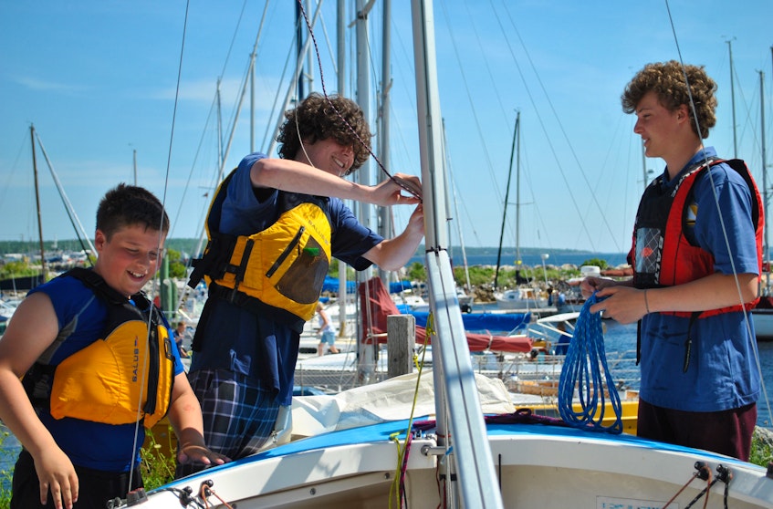 Shelburne Harbour Yacht Club Sailing Academy summer camp student Patrick Brannen (from left) helps instructors Rafael Pedro and Aaron Hobbs lower and store the sails on one of the Opti sailing dinghies   used to teach sailing at the academy. KATHY JOHNSON - Kathy Johnson