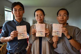 Edward Wang, left, his mom, Julie Zhu, and his dad, Jerry Wang, stand in their small, two-bedroom apartment in Charlottetown, holding their passports stamped "Cancelled Without Prejudice".