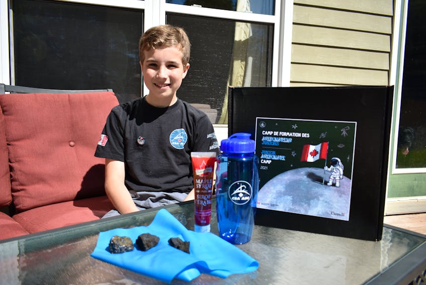 Brendan Reynard, 12, sits on his back deck in Charlottetown on Aug. 3, wearing a T-shirt and pin that were sent to him as part of the Canadian Space Agency's Junior Astronaut camp, which Brendan attended virtually July 26-30. On the table in front of him are some of the other goodies he received, including rocks commonly found on the moon, an exercise band and space maple syrup.
