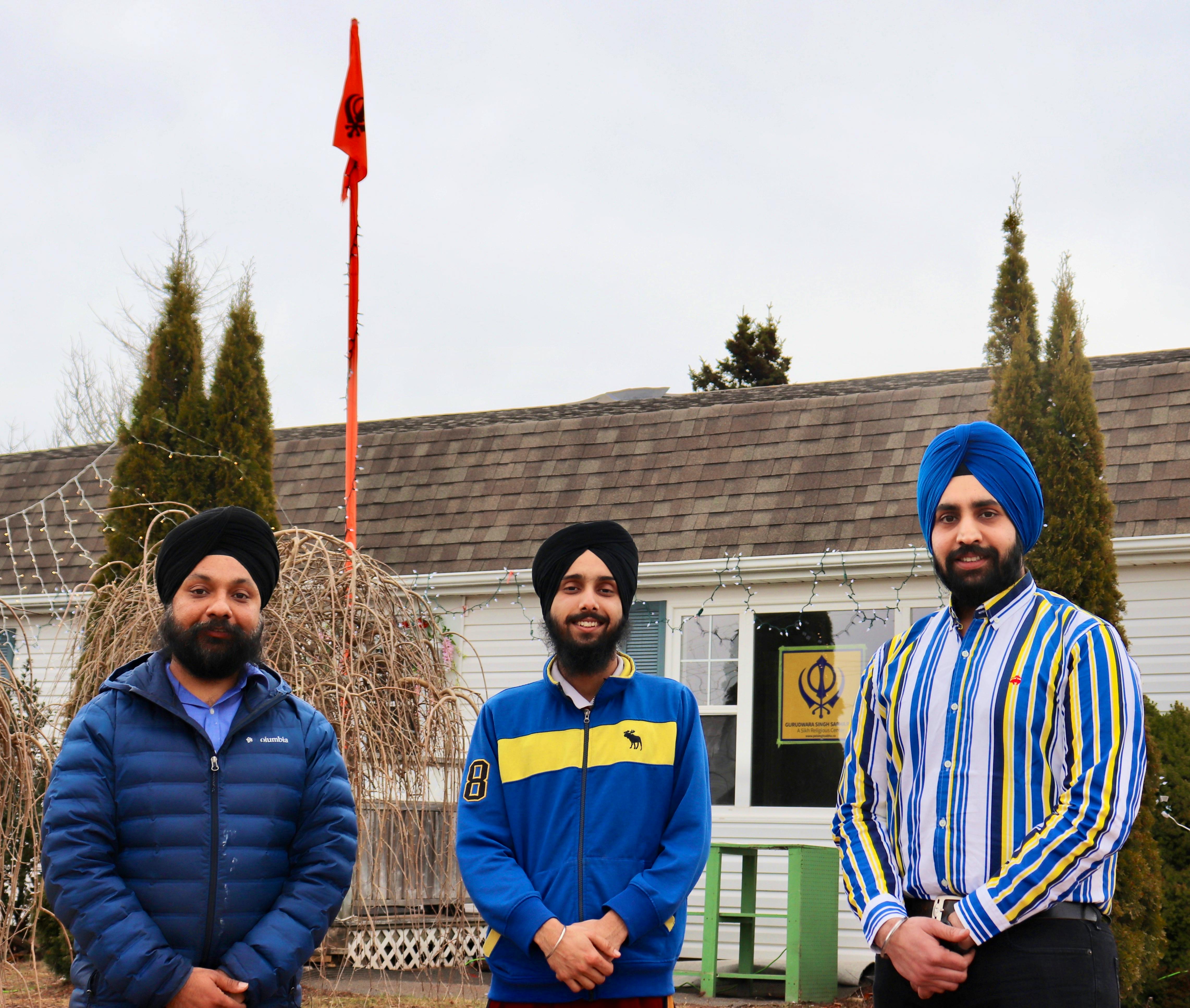 Prabhjot Singh, right, says gurudwaras across the world fly an orange flag (visibile in the background) called nishan sahib, which notifies people of a gurudwara in the neighborhood where they can eat and find shelter. Arshgod Singh stands in the middle with Savneet Singh on his left, outside the Gurudwara Sahib in Stratford.