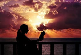 A woman reads a book outside by the water at sunset. - Pixabay