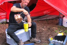 Darrell Hunt, sitting in front of his tent in a wooded area, pours hot water into his coffee cup. Contributed