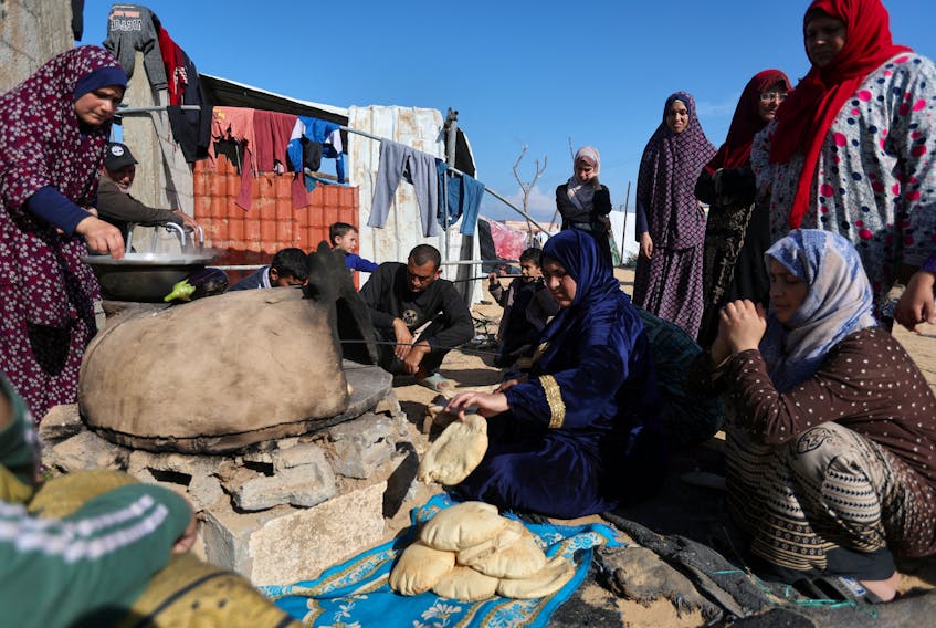 People wait while a woman prepares food, as displaced Palestinians, who fled their houses due to Israeli strike, shelter in a camp in Rafah, amid the ongoing conflict between Israel and Palestinian Islamist group Hamas, in the southern Gaza Strip, December 6, 2023.