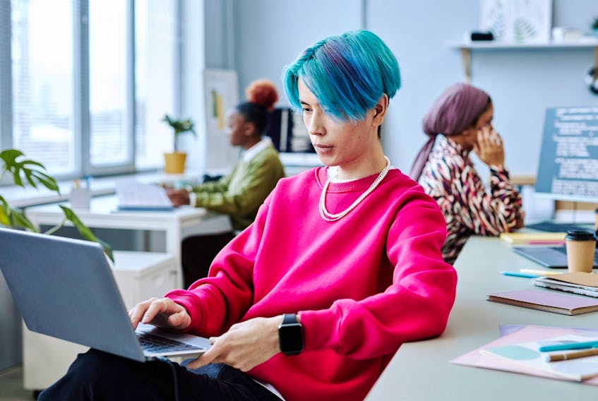 Young man with blue hair using laptop in modern office with two co-workers behind him.