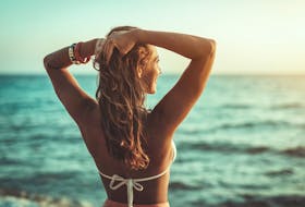 Back of young woman in bikini with raised arms enjoying sunset while looking out at ocean.