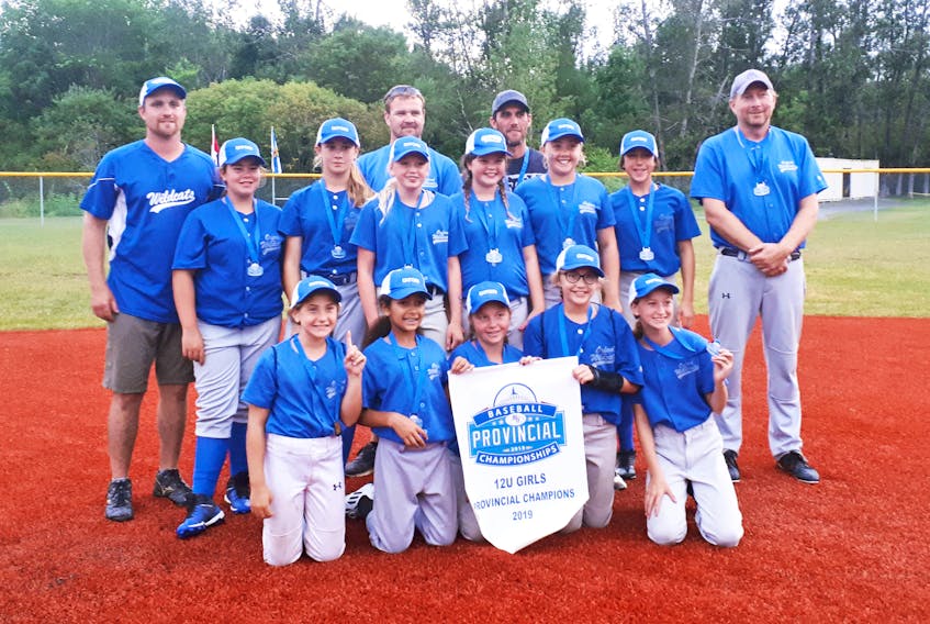 Wearing their championship medals, the Oxford Wildcats gather around the 12U provincial championship banner on the Sandlot baseball field in Antigonish. The winning team is: (front, from left) Julia Sharpe, Sierra Foley, Lily Dickey, Grace Weatherbee, Sophie Mazur, (middle, from left) Emily McLellan, Ella Tapper, Addie Fisher, Brooke Hunsley, Emma Reade, Allie Macdonald, (coaches, from left) Joe Reade, Ian Tapper, Blaine Weatherbee, and Jason Dickie.