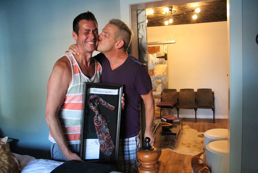 Ross Hunter, left, sneaks a kiss on his husband, Adrian MacKinnon’s, cheek. MacKinnon said he was in the closet for many years and still has a problem with being openly affectionate with Hunter, who he met in 2009. He is holding the “ugly tie” he was wearing when they met because Hunter used it as a way to introduce himself. The couple framed it with their wedding invitation.