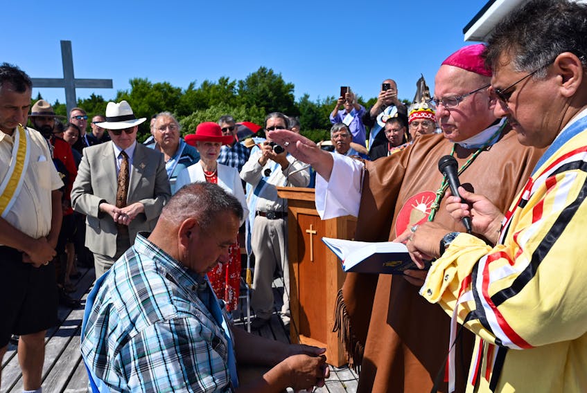 New Mi’kmaq Grand Chief Norman Sylliboy of Eskasoni (kneeling) receives a blessing from Bishop Brian Dunn while diaconate candidate Keptin Thomas Sylliboy holds a mic at the end of mass during the annual Mission of St. Anne on the island near Potlotek on July 28. Grand Chief Sylliboy is the grandson of the late Grand Chief Gabriel Sylliboy of Eskasoni. Among those looking on, from front left, are Grand Keptin Andrew Denny, Lieutenant Governor Arthur LeBlanc and his wife Patsy LeBlanc.
