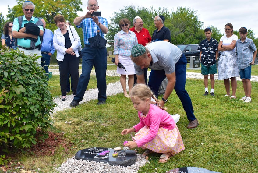 Five-year-old Ila Simon places some specially decorated stones on a plaque honouring her late grandfather, Dr. Phil Simon, who was remembered during a special ceremony at Glace Bay’s Hebrew cemetery on Sunday that also recognized the late Elliot (Ellie) Marshall. Friends and relatives look on as Ila’s father Mark, bent over and wearing a Cape Breton tartan kippa or yarmulke, gets another stone ready for his daughter. He collected the stones in Israel.