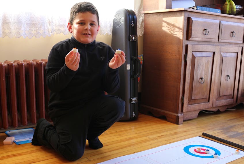Colton McVarish, 7, kneels next to his practice curling game holding up miniature stones at his home in New Victoria on Wednesday. McVarish has earned the No. 5 spot in the Hit Draw Tap curling competition for the province of Nova Scotia.