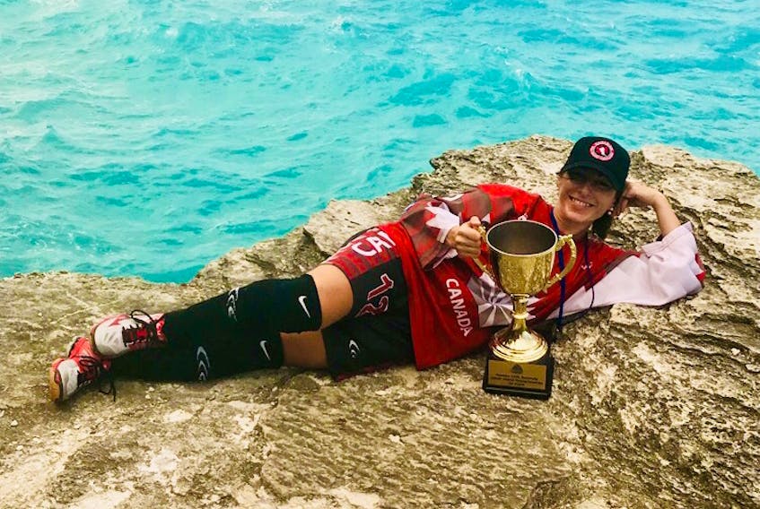 Susan Stuckless in her red Team Canada jersey displays her gold medal and the team trophy near the ocean in Bermuda. Stuckless says since the ocean was so close to the rink, players would often jump into the water to cool off after games.