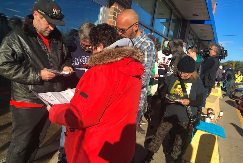 An estimated 100 people wait in line at the Nova Scotia Liquor Commission outlet in Sydney River to purchase legal cannabis. They are shown reading over information on various products.