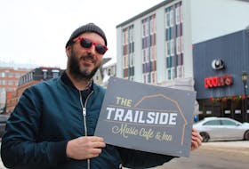 Pat Deighan holds an old business sign for The Trailside Music Café and Inn, which is moving out of Mount Stewart and into the tall, gray building behind him as Trailside Music Hall next year.