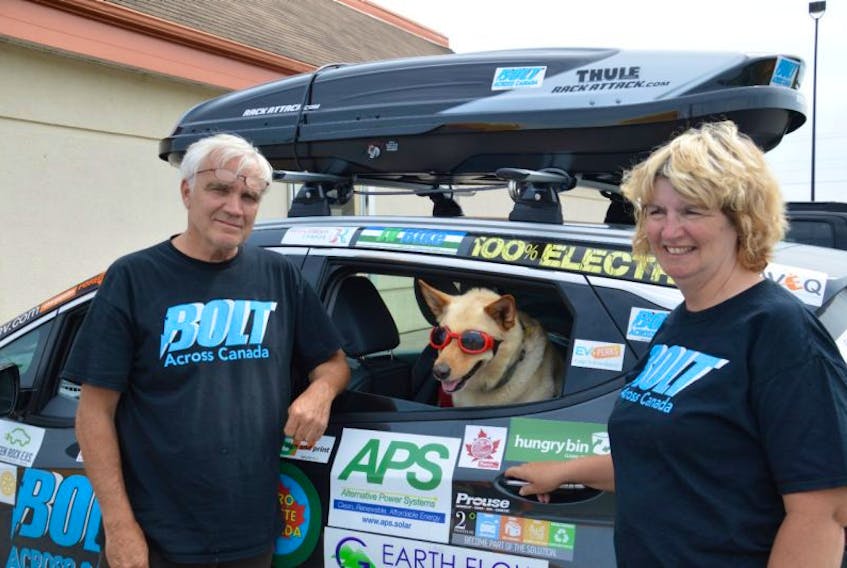 Buddy Boyd and his partner, Barbara Hetherington, stand beside their Chevy Bolt electric car while their rescue dog, Piper, shows off her driving goggles.