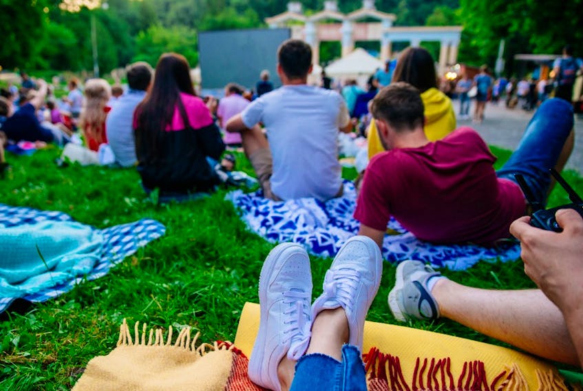 People lay sprawled with their friends on picnic blankets at a park as they watch an outdoor movie. 